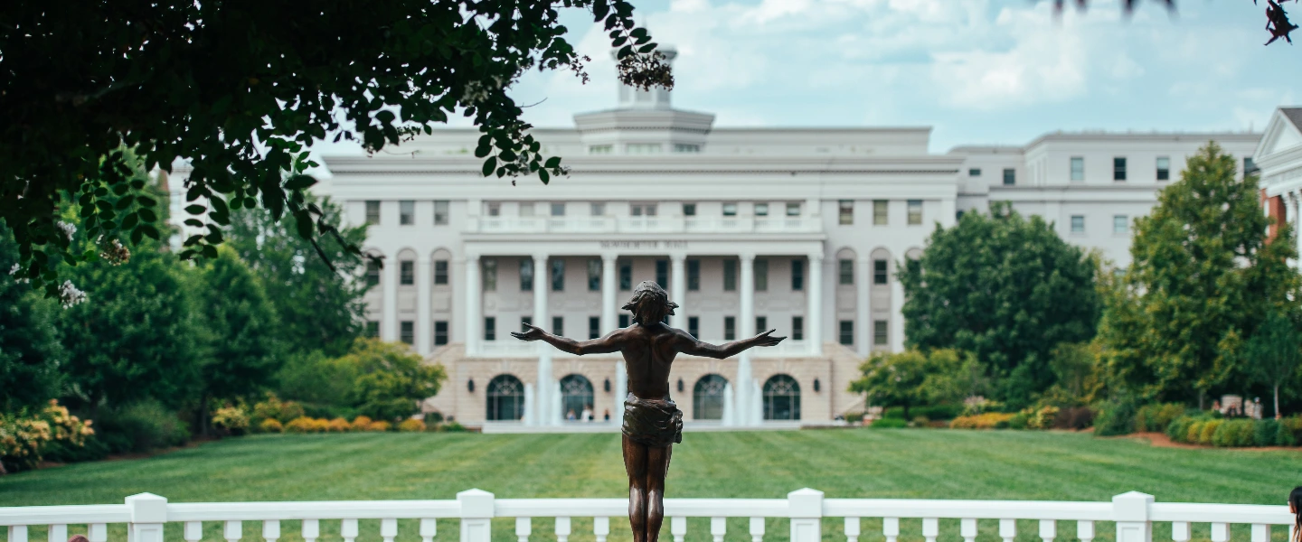 Bronze statue with open arms facing a grand white collegiate building with pillars and a lush green lawn.