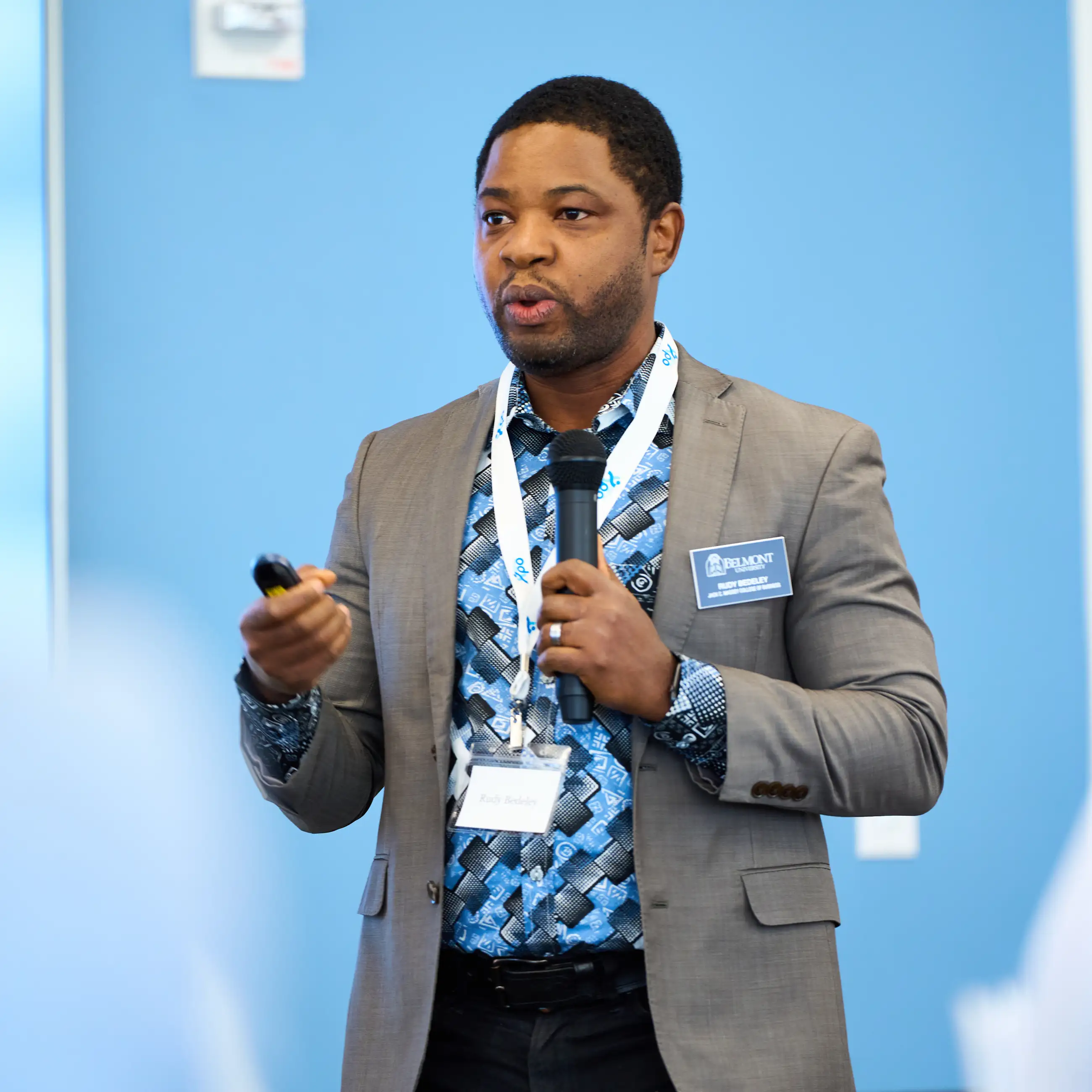 A Belmont University faculty member speaks during a presentation, holding a microphone and clicker while standing against a blue background.