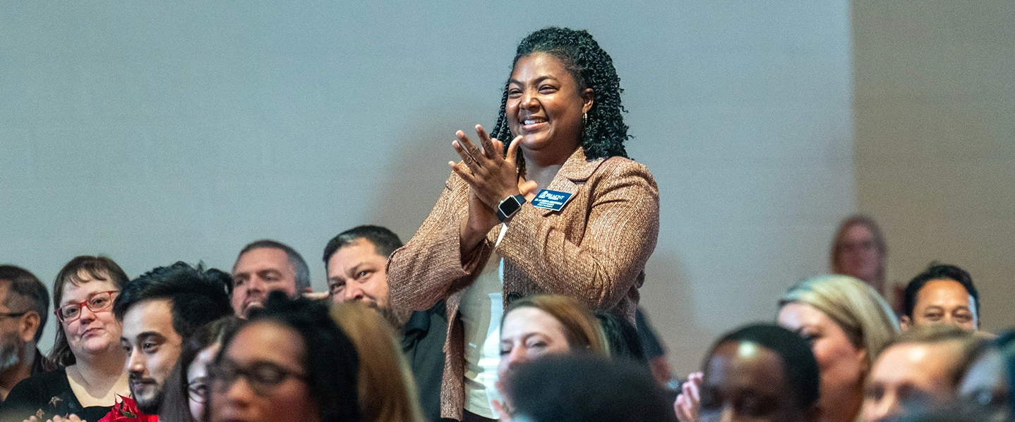 A Belmont employee stands up at a staff gathering and is clapping.