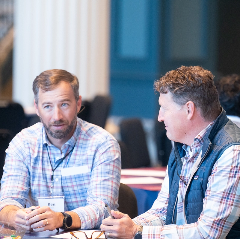 wo men sit at a table, engaged in conversation. One is wearing a plaid shirt and a name tag reading "Ben," while the other wears a vest and listens attentively.