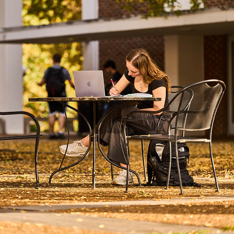 A student sitting a a table outside writing in a notebook with her laptop in front of her