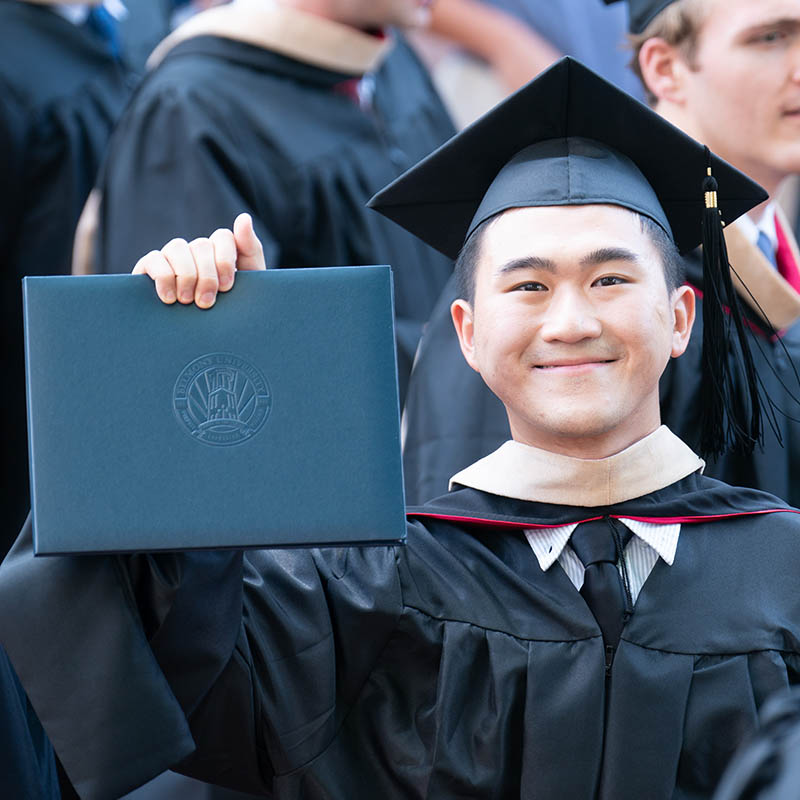 A graduate student in a cap gown and hood holding up his diploma after graduation