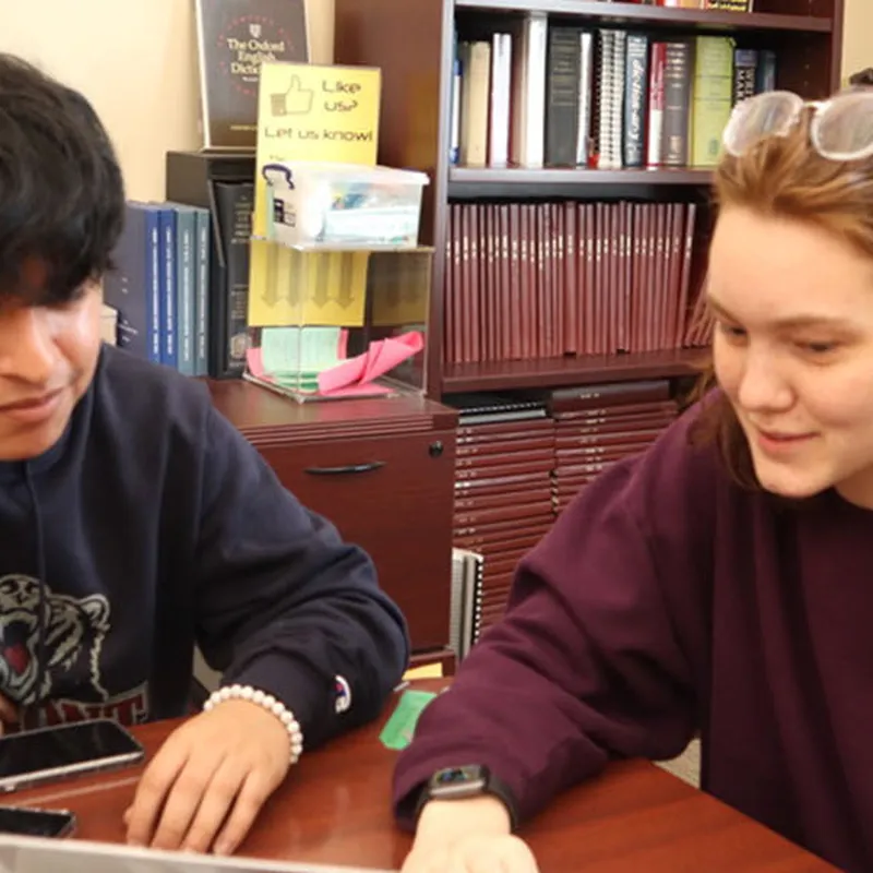 Two students collaborating at a table in a writing center, reviewing work together with bookshelves and study materials in the background.