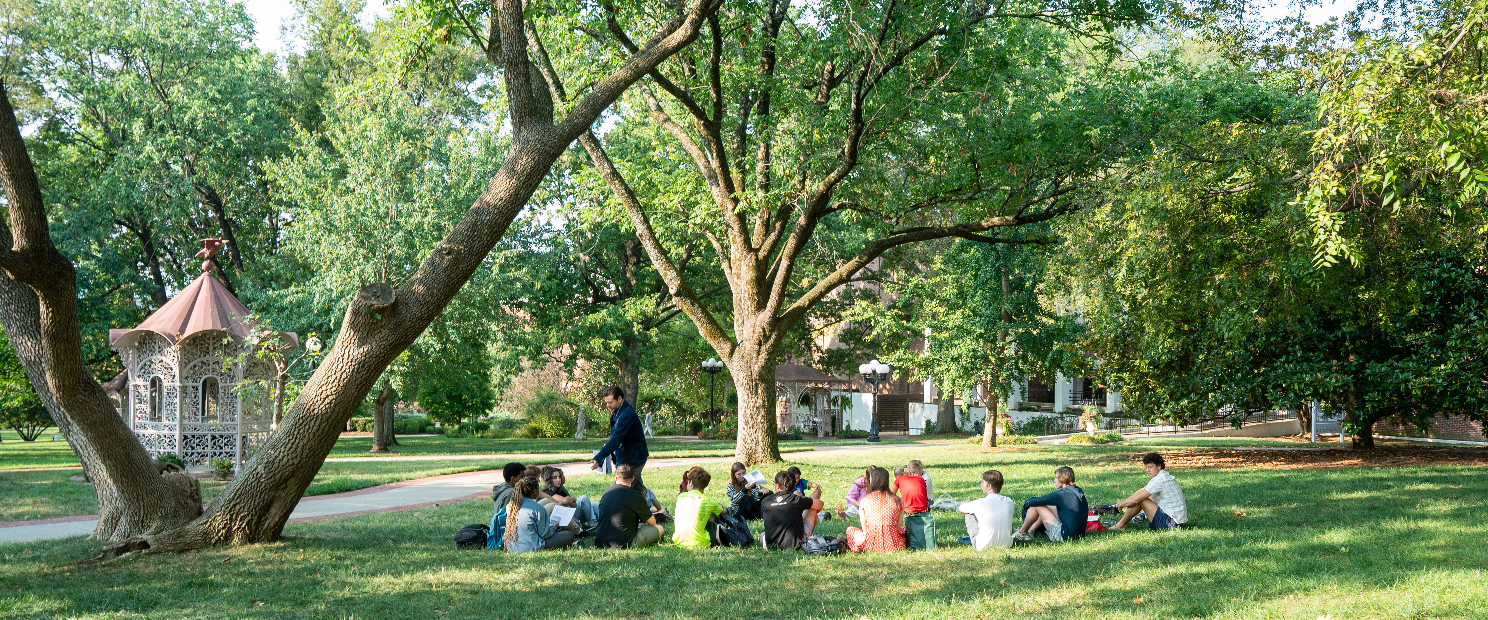 Students sitting on the lawn on the historic side of campus under trees during class outside