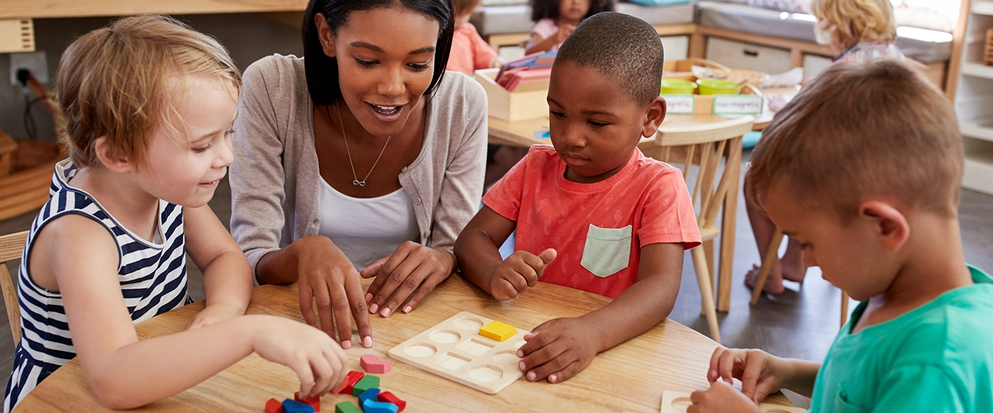 A young woman at a table showing blocks to preschoolers