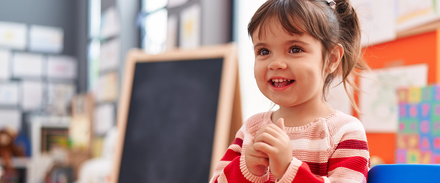 A little girl in a striped sweater clasps her hands and smiles in a classroom with an easel chalkboard behind her