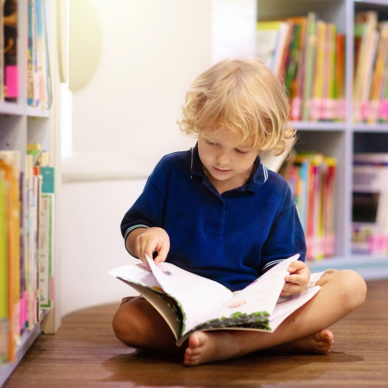 A little boy is seen seated in front of two bookshelves with an open book in his lap.