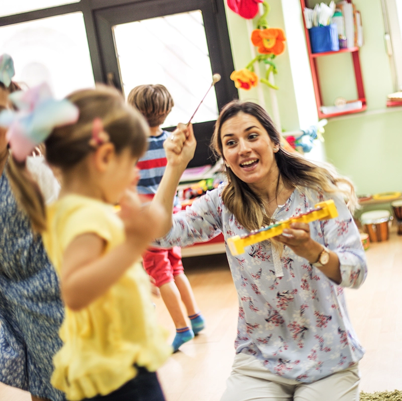 A young teacher playing a xylophone while children dance around her.