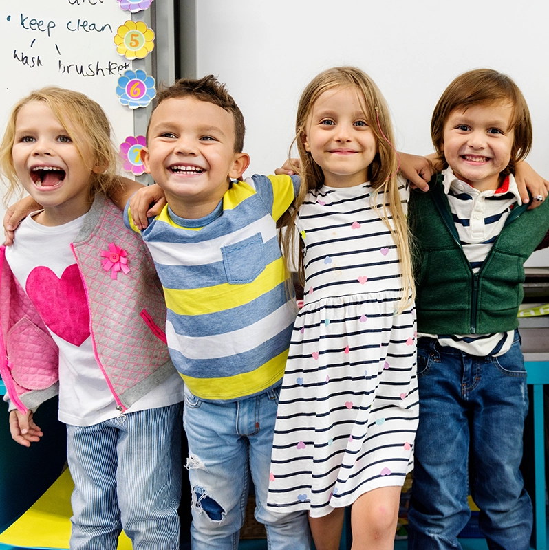 Preschoolers with their arms around each other standing in front of a classroom and smiling