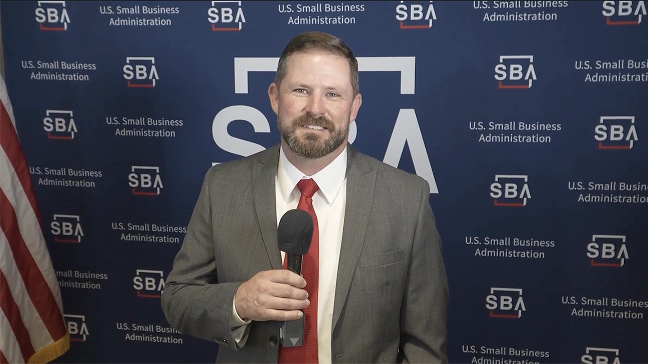A business man speaking with a microphone in front of a Small Business Week Background
