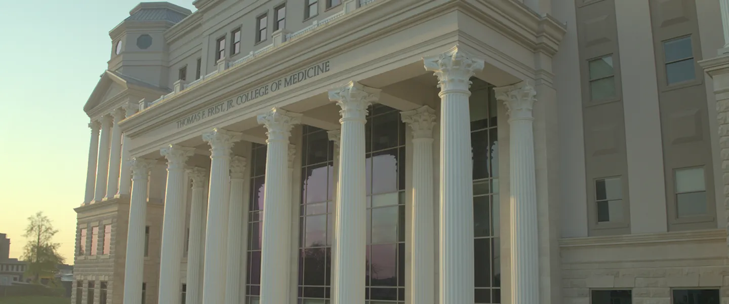 A close-up view of the front entrance of the Thomas F. Frist, Jr. College of Medicine at Belmont University. The building features grand, white Corinthian columns that line the entrance, supporting a detailed entablature with the college's name engraved across the top. Large windows between the columns reflect the soft light of the setting sun, casting a warm glow over the impressive classical architecture. The surrounding stonework and design elements emphasize the building's grandeur and institutional significance.