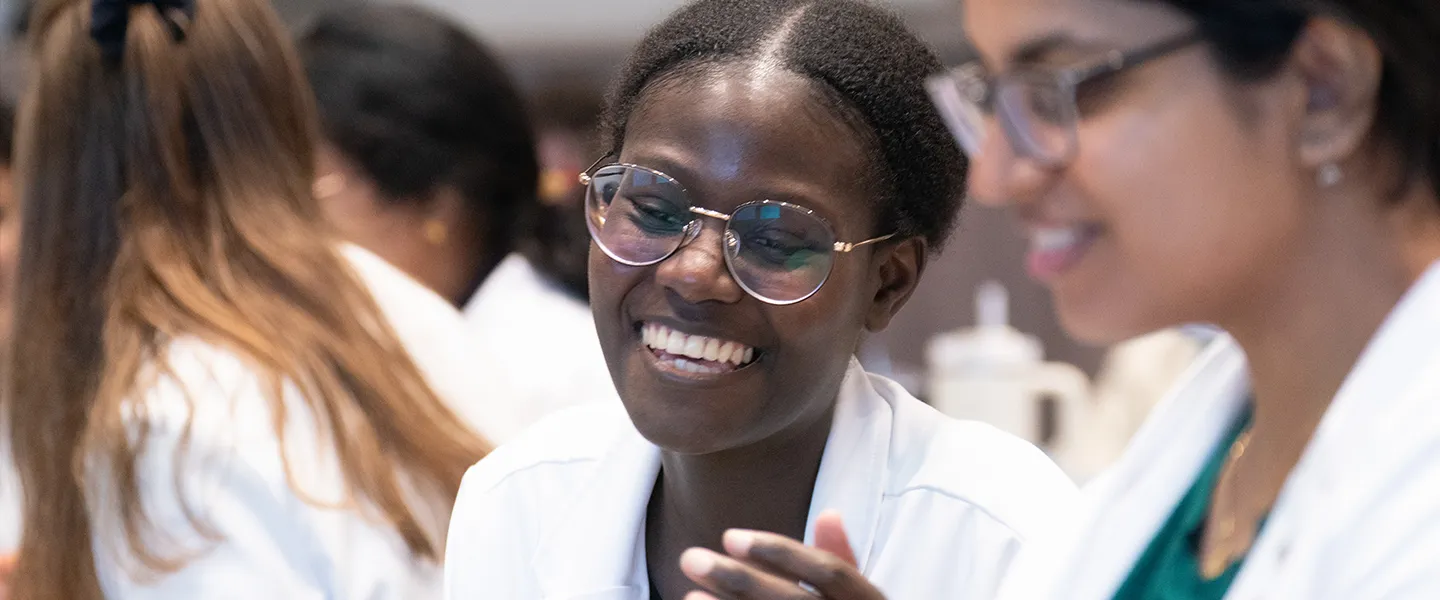 Two medical students are seen smiling and engaging in conversation, wearing white lab coats in what appears to be a collaborative academic setting. The student in the foreground has round glasses and a bright, joyful expression, while the other student, slightly out of focus, is also smiling and wearing glasses. The background is filled with other students, suggesting a lively and interactive classroom or laboratory environment.