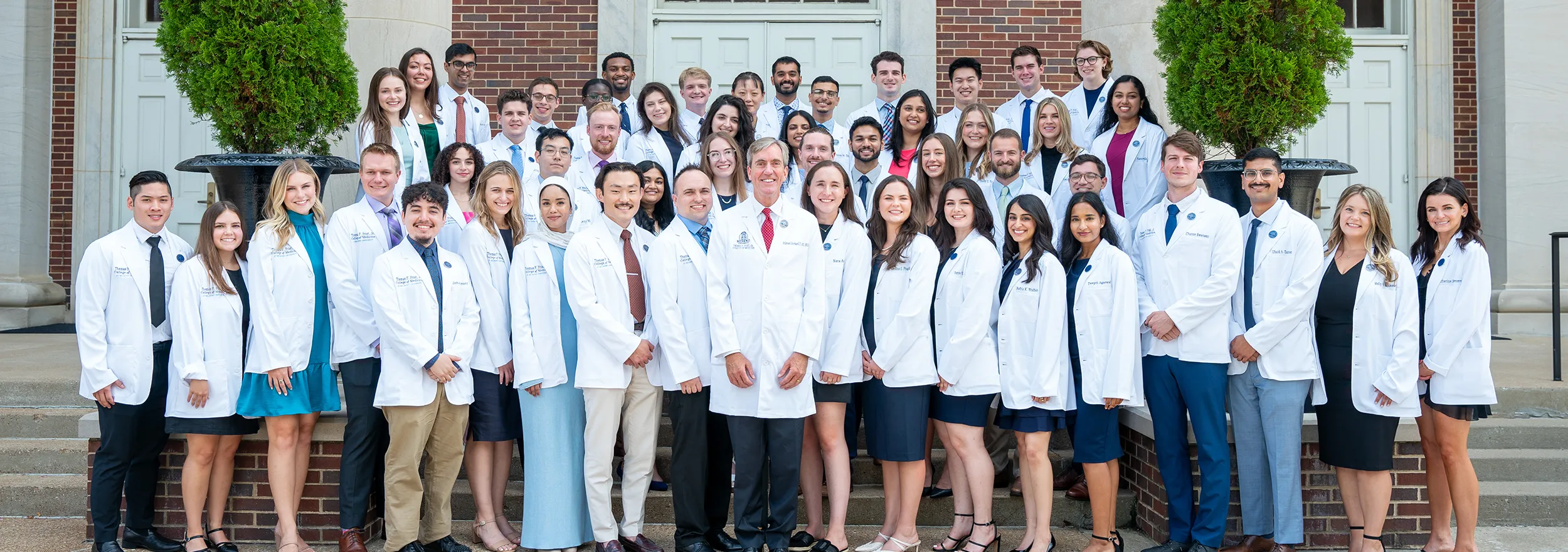 The inaugural class of the Thomas F. Frist, Jr. College of Medicine at Belmont University poses for a group photo on the steps of a campus building. The students, wearing white lab coats, stand together smiling and looking towards the camera. 