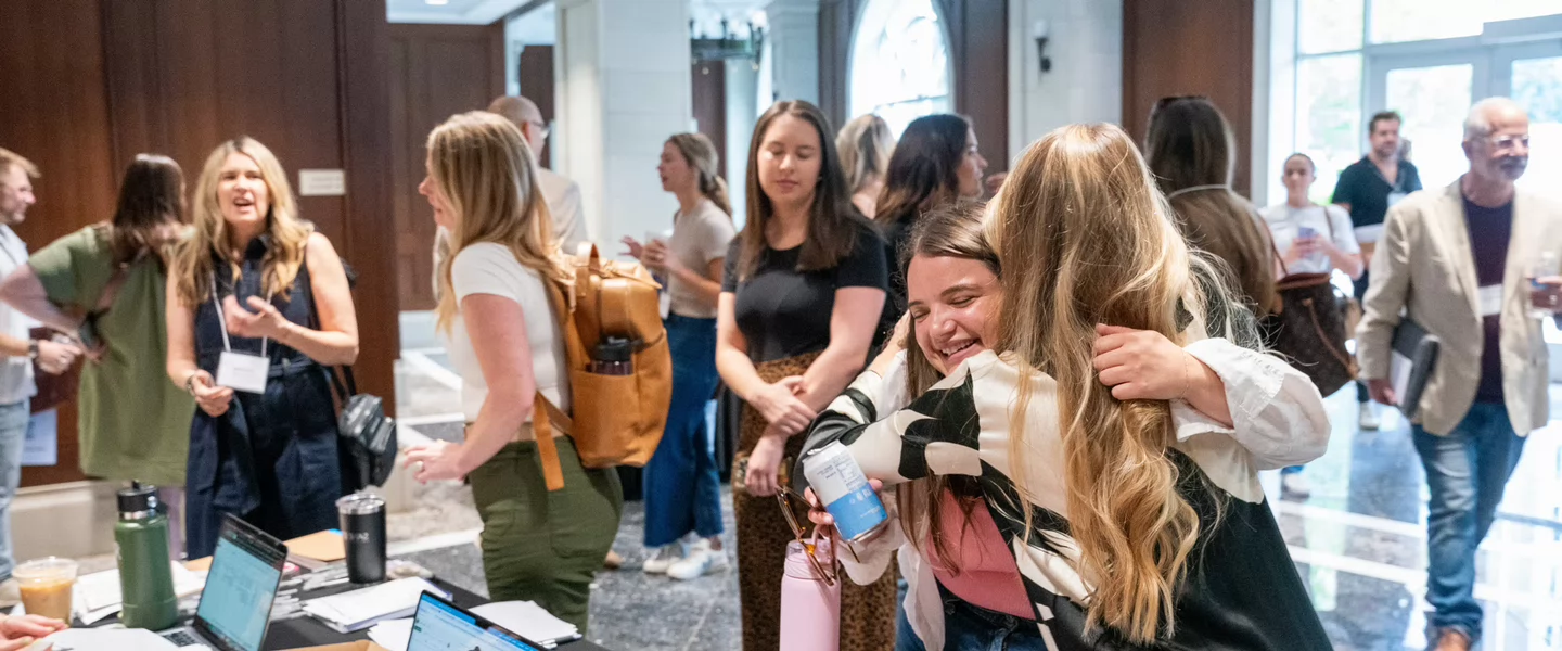 Attendees greet and hug one another during a networking gathering at Belmont University’s 24/7 Conference, with conversations and check-in tables in the background.