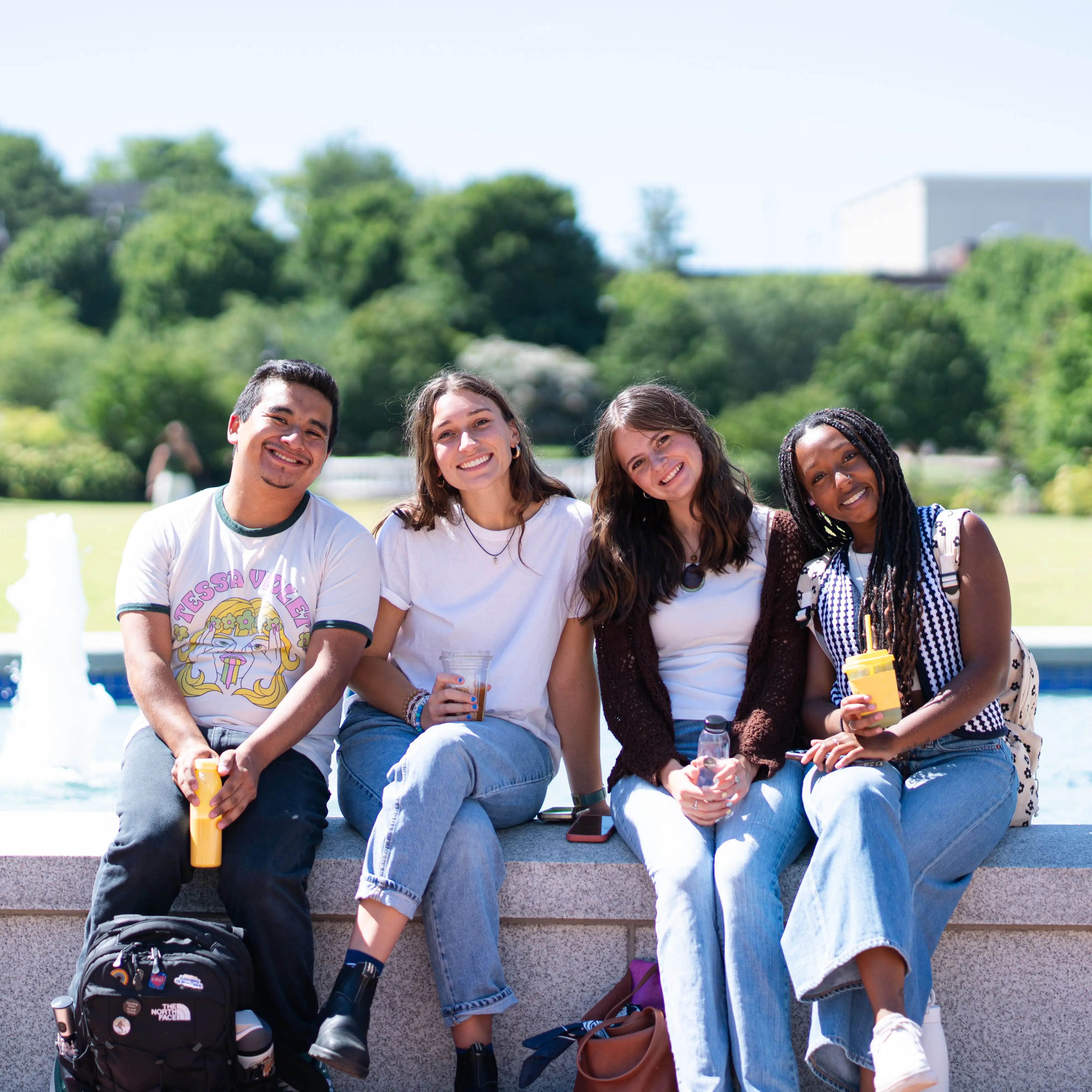 Four students sit together on a low stone wall near a campus fountain, smiling and holding drinks on a sunny day at Belmont University.