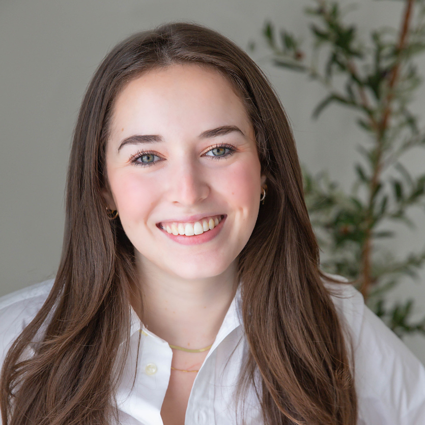 Professional headshot of Ansley Deaton, featuring her with long straight brown hair, blue eyes, and a bright smile. She is wearing a white button-down shirt and gold jewelry, posed against a light background with greenery in soft focus.
