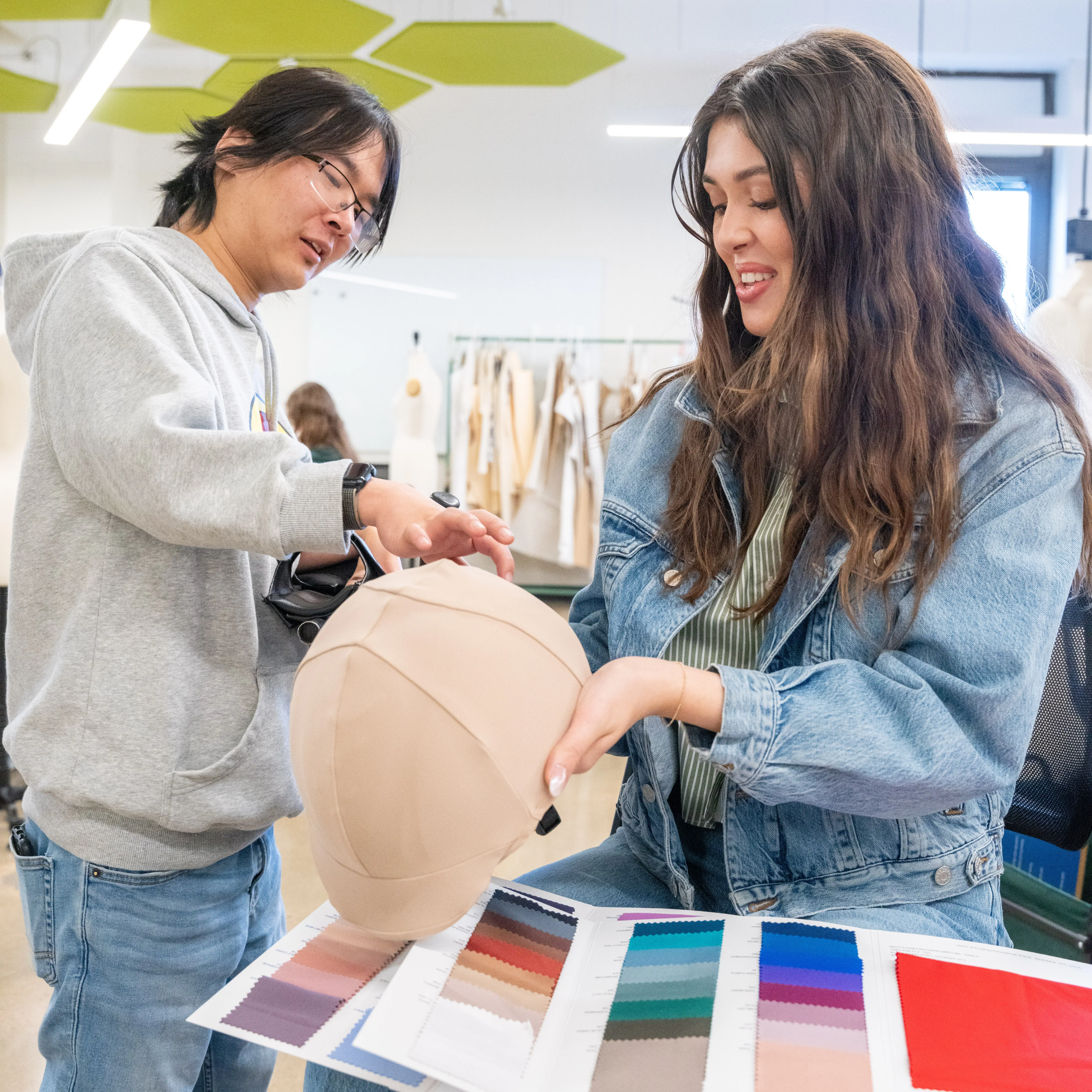 Two Belmont University fashion students collaborate in a design studio, reviewing fabric swatches and examining a hat prototype during a project discussion.