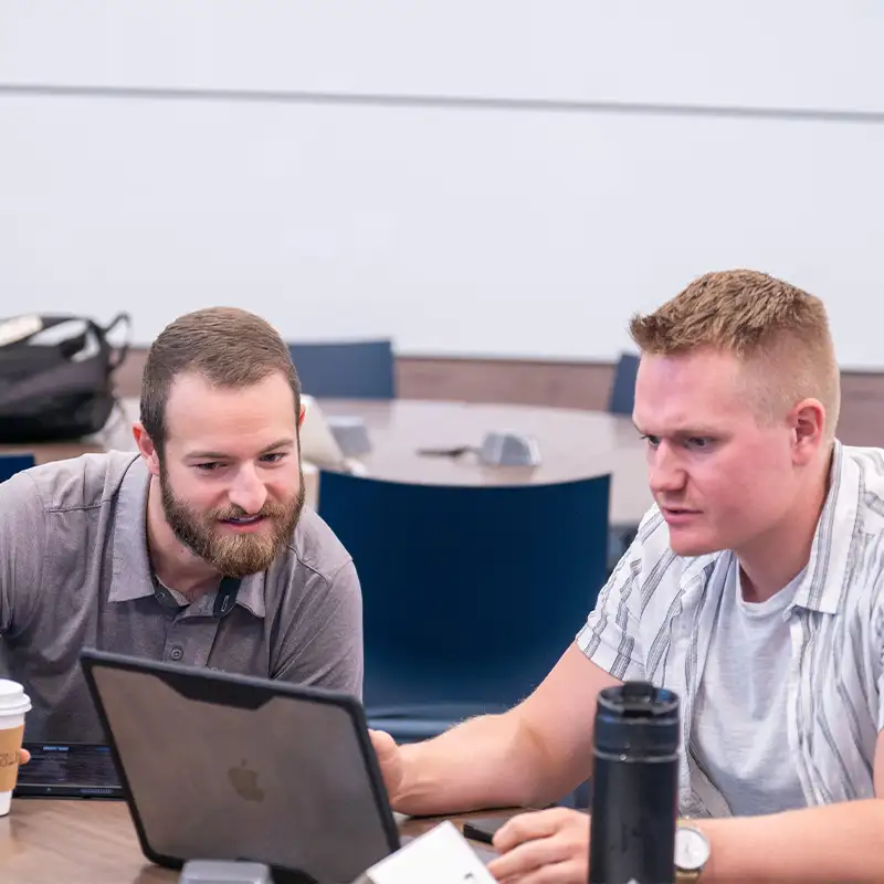 An indoor photo of two men seated next to each other and looking at a laptop together