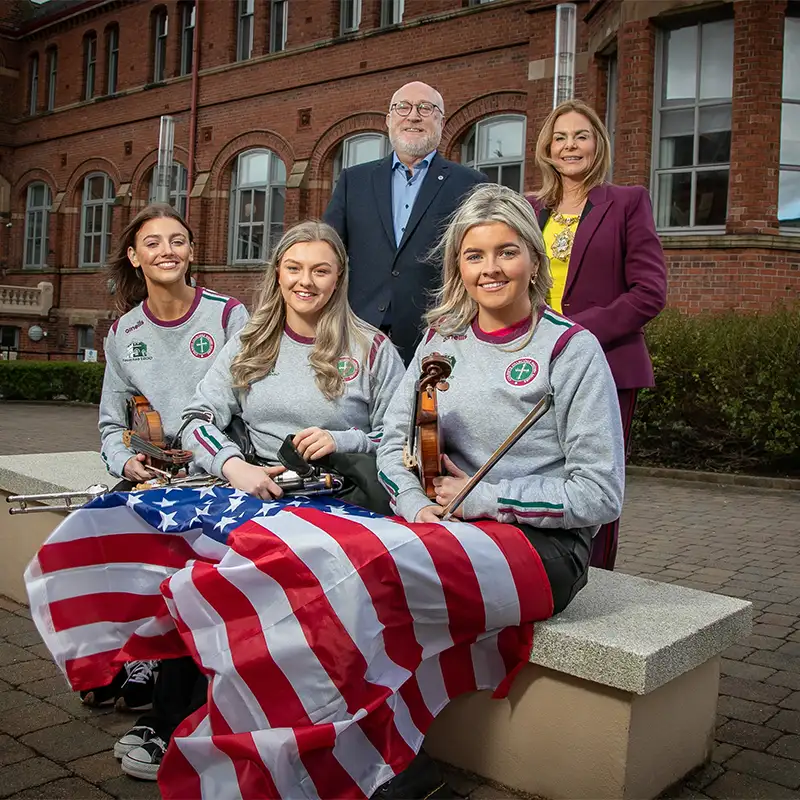 An outdoor photo of 3 female Belfast students seated with 2 faculty members standing behind them with a United States flag draped over their legs while holding instruments