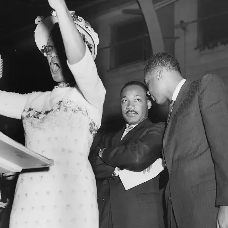 An indoor photo of a woman speaking at lectern in foreground, while Dr. Martin Luther King stands behind and man is speaking to him. 