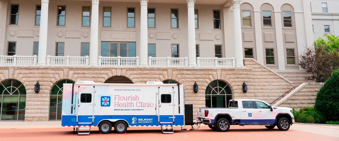 A Belmont University mobile clinic, labeled “Flourish Health Clinic,” is parked in front of a campus building with white columns. A red ribbon is tied across the front of the trailer, and a woman in a blue and white dress speaks at a podium next to a Belmont-branded truck.