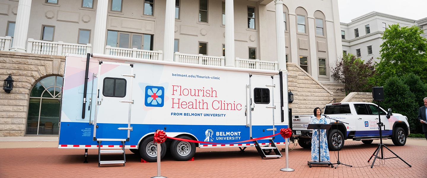 A Belmont University mobile clinic, labeled “Flourish Health Clinic,” is parked in front of a campus building with white columns. A red ribbon is tied across the front of the trailer, and a woman in a blue and white dress speaks at a podium next to a Belmont-branded truck.