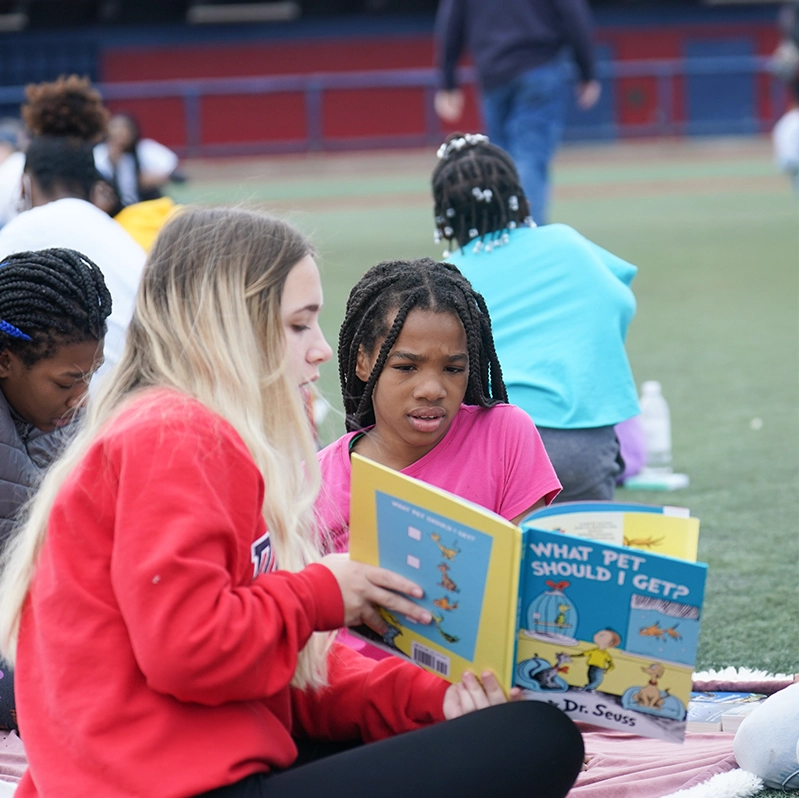 A college student in a red sweatshirt reads a Dr. Seuss book titled What Pet Should I Get? with a young girl sitting beside her on a field, surrounded by other children reading.