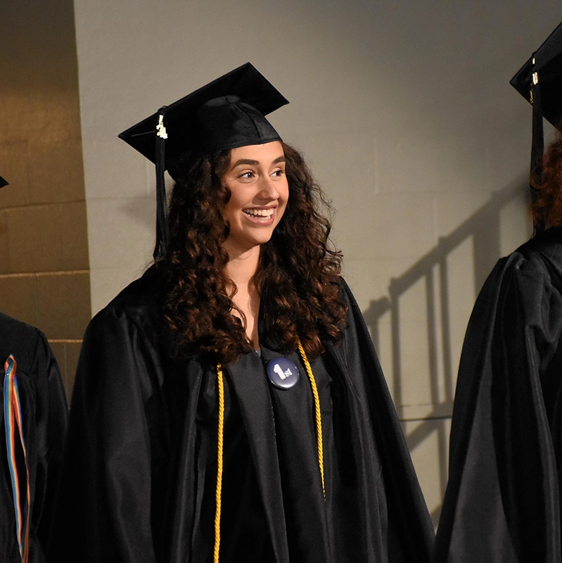 A smiling graduate wearing a black cap and gown stands in line during a commencement ceremony, shown with long curly hair and an honor cord draped around her neck.