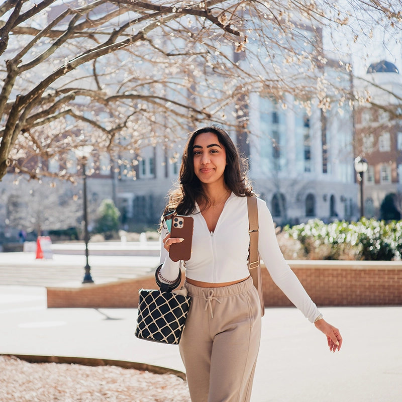 Belmont University student walking on campus, holding a phone and smiling, with academic buildings in the background.