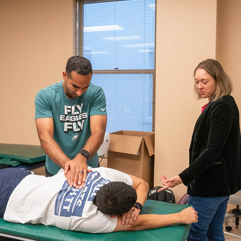 Three people engaged in a physical therapy session: one individual is lying face down on a treatment table while another performs a manual therapy technique on their back, supervised by a third person who is observing and providing guidance.