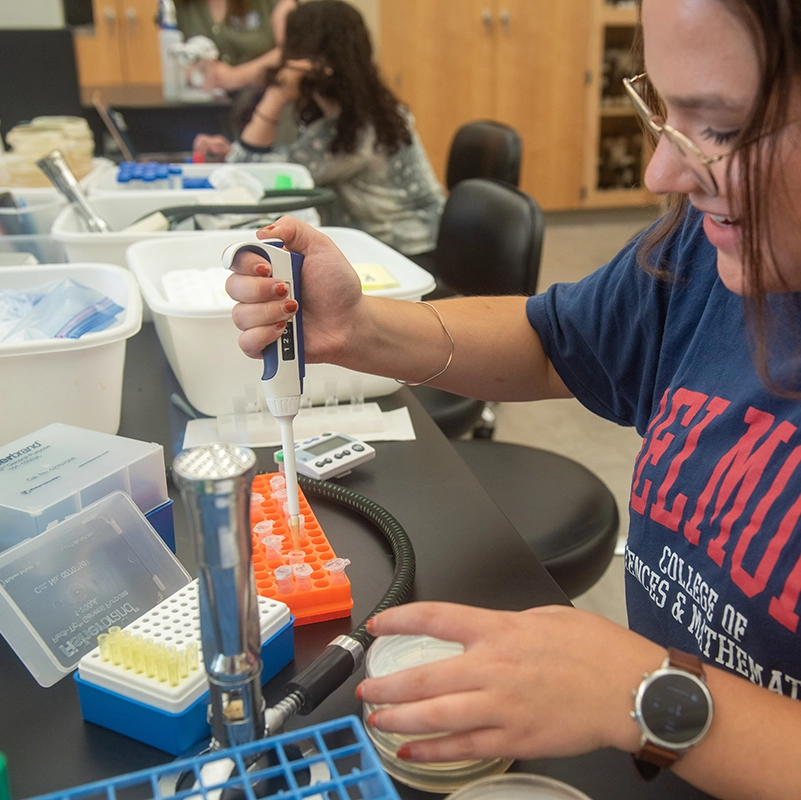  Student in a Belmont College of Sciences and Mathematics T-shirt uses a pipette in a laboratory, working with test tubes and lab equipment on a table.