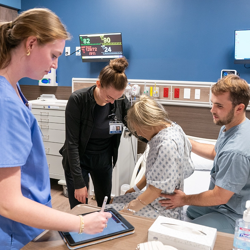 Belmont University healthcare students working together in a clinical simulation, assisting a patient and recording data on a tablet.