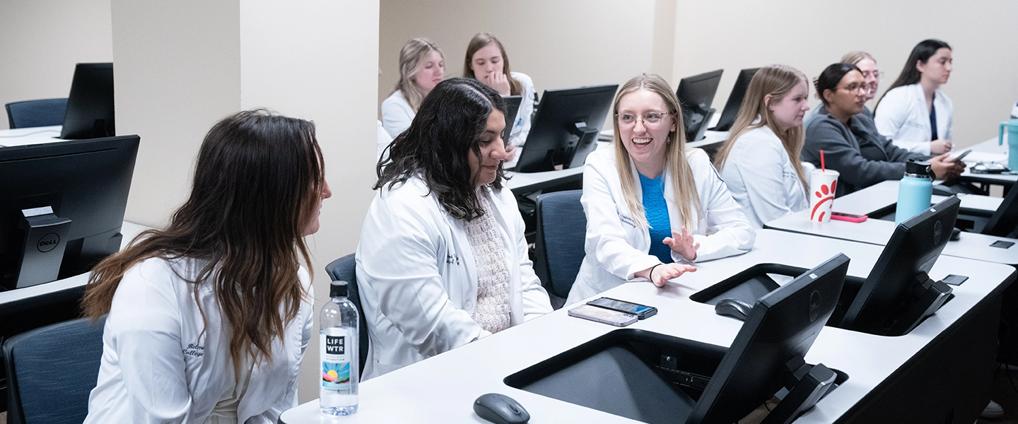 A group of students in white lab coats engage in a discussion in a classroom with computer workstations, reflecting a collaborative learning environment in a healthcare education setting.