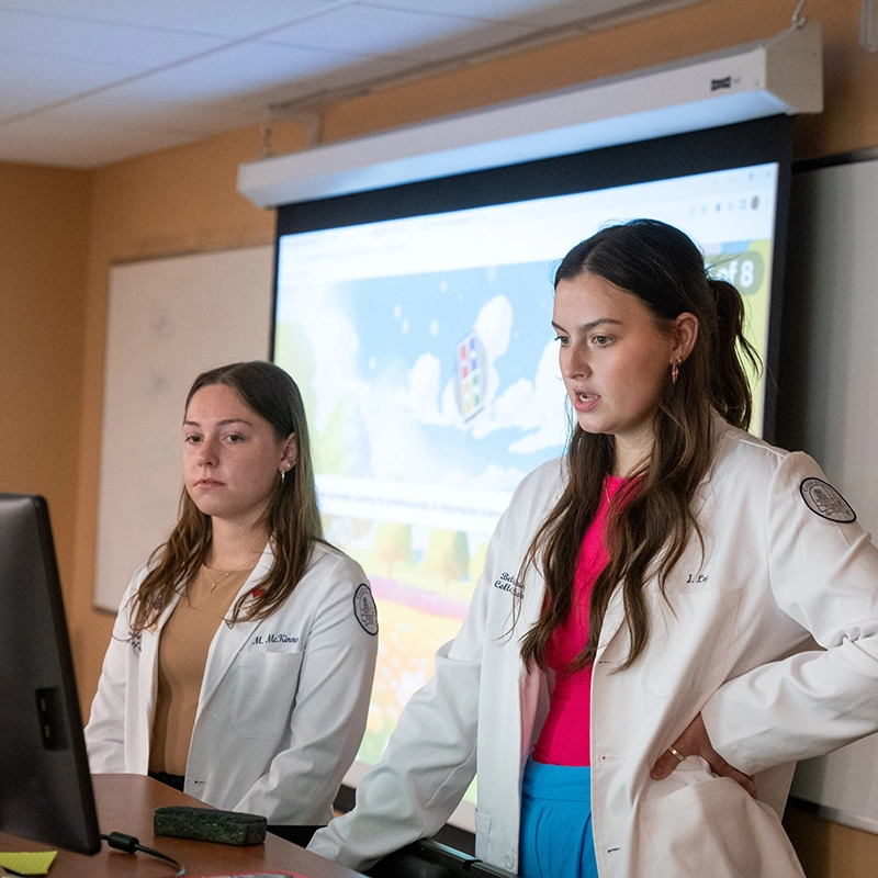 Two students in white lab coats present in front of a classroom with a digital screen displaying colorful graphics, showcasing a professional and educational setting in health care learning.