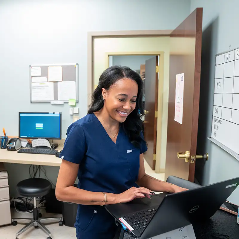 Woman in scrubs at doctors office smiling while typing on a computer