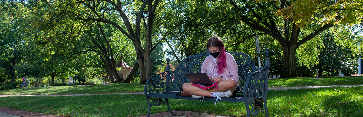 A student in a mask on a bench working on a computer