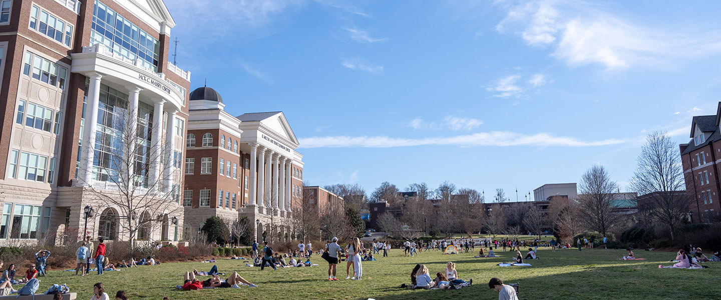 Students relax, study, and socialize on a large grassy lawn under a sunny blue sky in front of Belmont University's Jack C. Massey Center and nearby academic buildings.