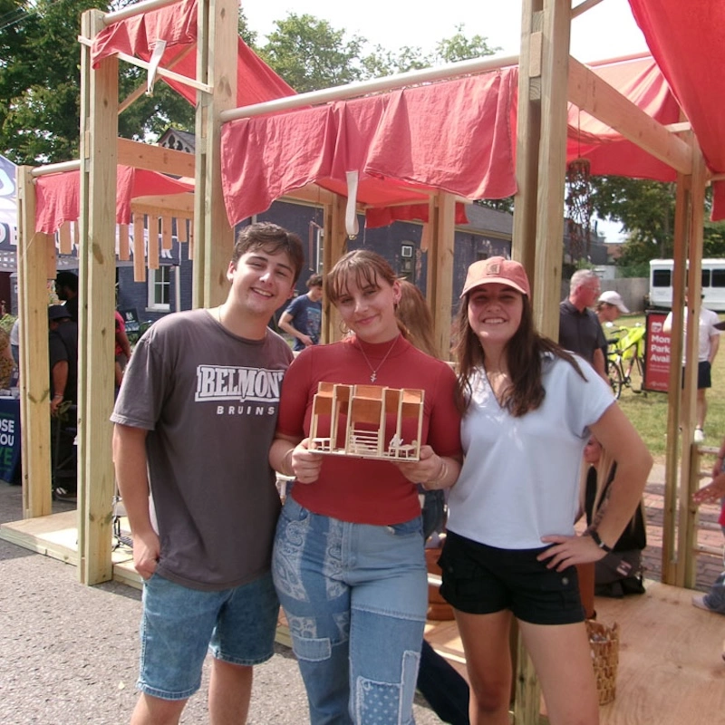 Three Belmont University students pose proudly at the PARKing Day event, standing in front of their design installation. One student holds a model of their project, while the others smile beside her. The backdrop features colorful canopies and a lively atmosphere, celebrating their achievement in the design competition.