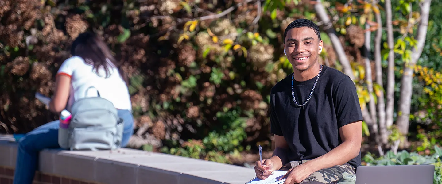 A student sits outdoors on Belmont University's campus, smiling while holding a pen and notebook. Another student studies in the background, surrounded by trees and autumn foliage.