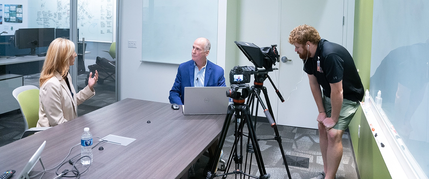 A man and woman seated adjacent from each other at a rectangular desk while a photographer is manning a camera