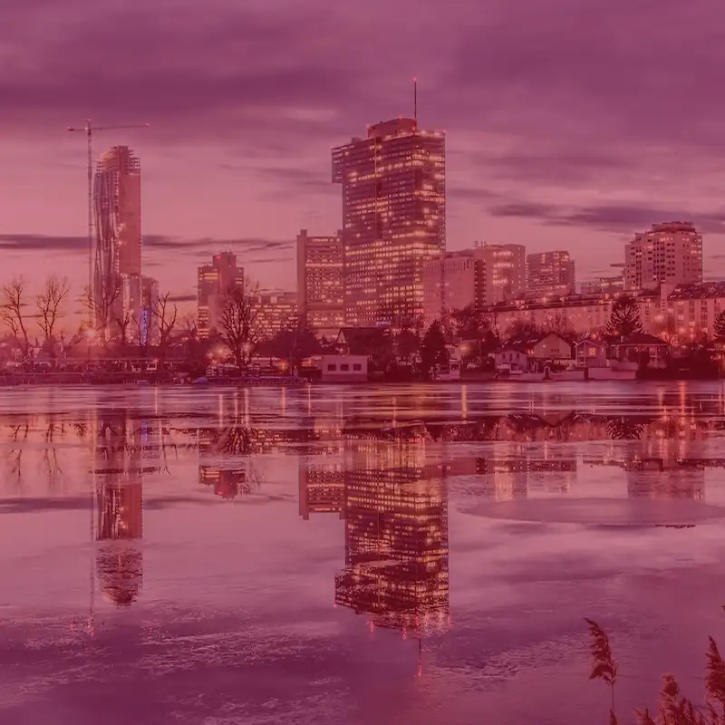 A city skyline at dusk with illuminated skyscrapers and smaller buildings reflected in the calm waters of a river in the foreground.
