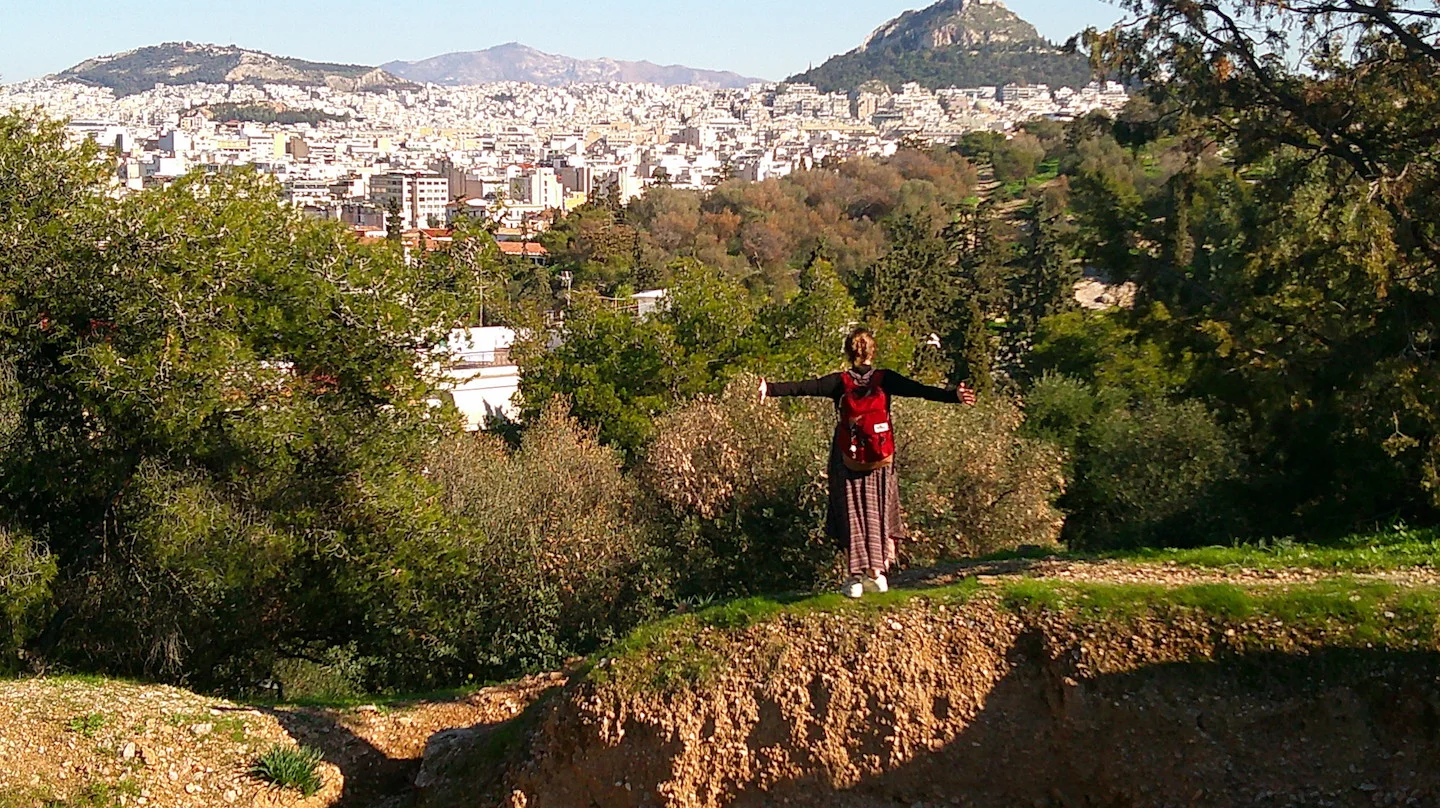 A student with arms outstretched enjoying a panoramic view of Athens, Greece from a hillside lookout point. The expansive cityscape stretches to distant mountains under bright sunshine.