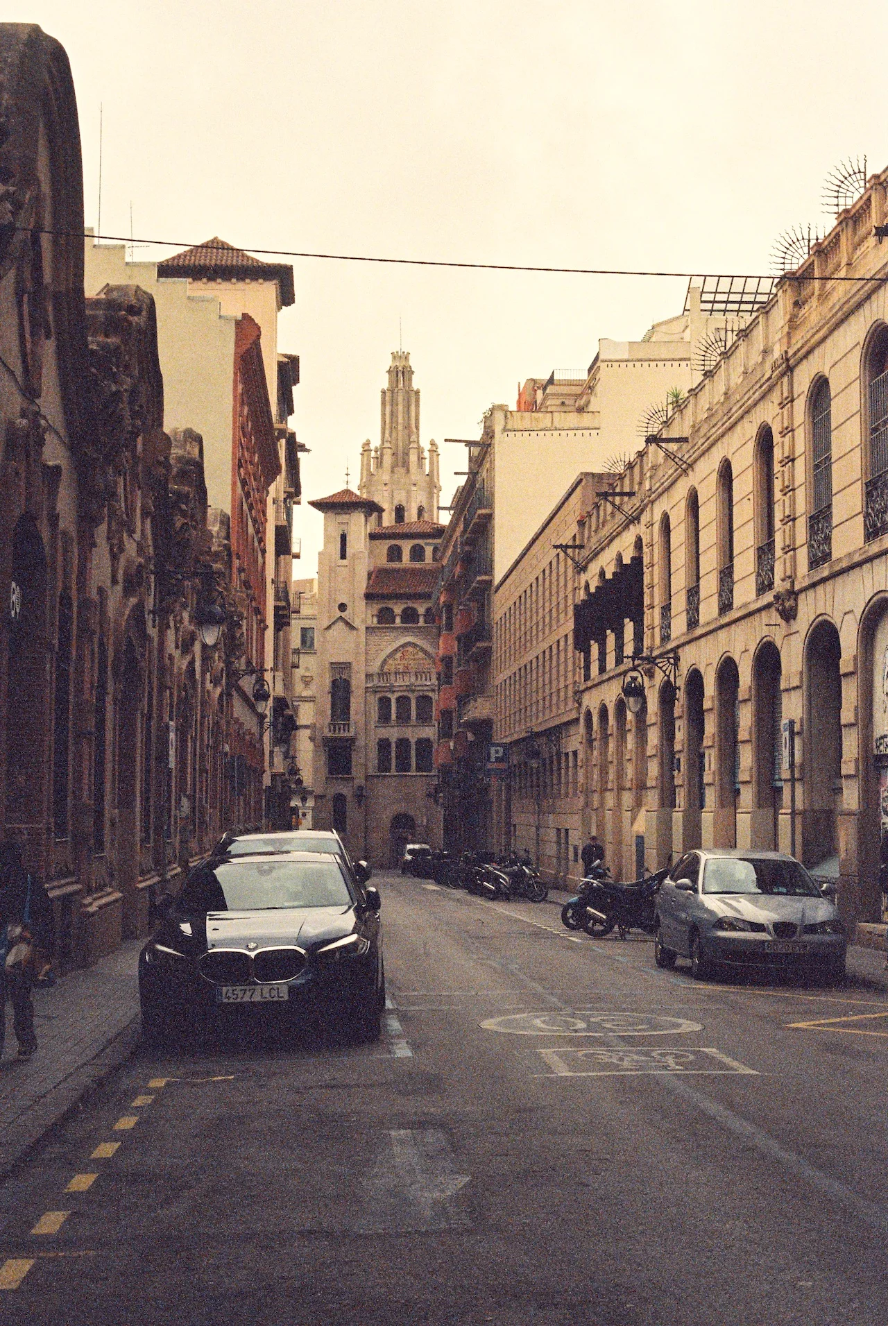 The impressive Plaza de España complex in Seville, Spain, showcasing its Renaissance and Moorish revival architecture bathed in warm sunlight. The Spanish flag flies prominently from one of the towers. The building features ornate stonework, arched galleries, a bell tower with blue-tiled dome, and intricate decorative elements against a clear blue sky.