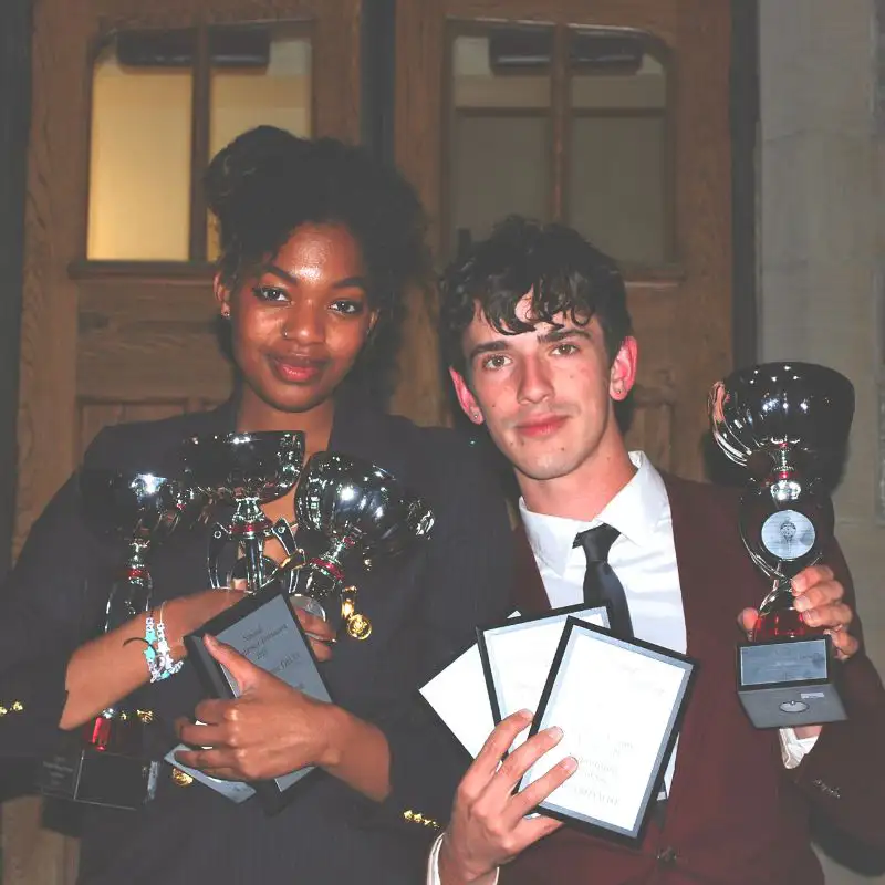 A young woman and a young man standing side by side in front of a set of wooden doors, each holding multiple shiny trophies and award plaques. The woman on the left smiles and cradles several silver cups and framed certificates in her arms, while the man on the right holds a large trophy in one hand and three plaques in the other. Both are dressed in formal attire and appear proud of their accomplishments.