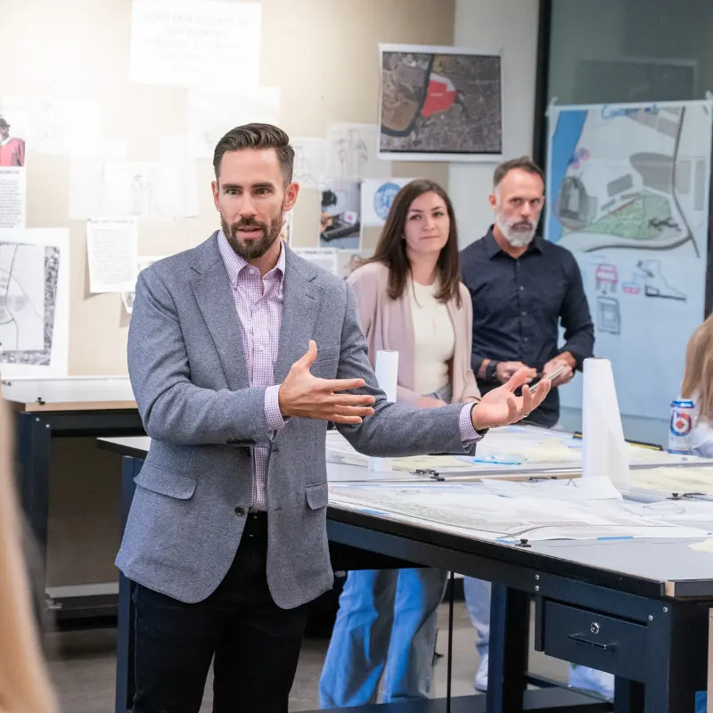 A man with a beard is standing in a classroom setting, gesturing as he speaks. He is wearing a light gray blazer over a checkered shirt. Two other individuals are visible in the background: a woman with long hair dressed in a light sweater and a man with a beard in a dark shirt. Behind them, various architectural drawings and plans are pinned to the wall, indicating a design or planning discussion is taking place. The atmosphere appears collaborative and focused.