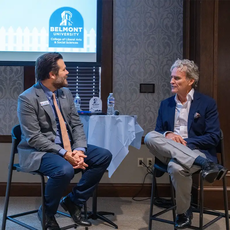 Two speakers engage in a discussion during a symposium at Belmont University. One speaker, dressed in a gray blazer and orange tie, listens attentively while the other, in a blue suit, shares insights. A table with water bottles and a Belmont University sign is visible in the background, creating a professional atmosphere for the event.