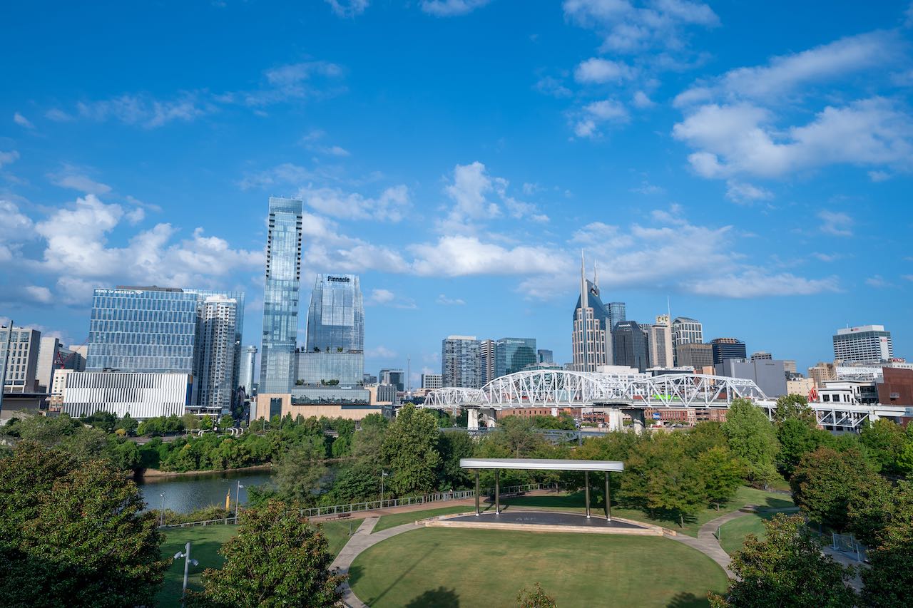 Nashville skyline on a sunny day, featuring modern skyscrapers, a white truss bridge, and a park with green trees in the foreground. The scene includes the Cumberland River and a clear blue sky with scattered clouds.