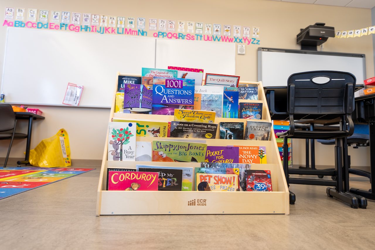 Close-up of a classroom reading corner. A wooden book display stand is filled with various children's books. Above it, the wall displays a complete alphabet with colorful letters and corresponding flashcards. A whiteboard and projector are visible, along with part of an educational rug on the floor.