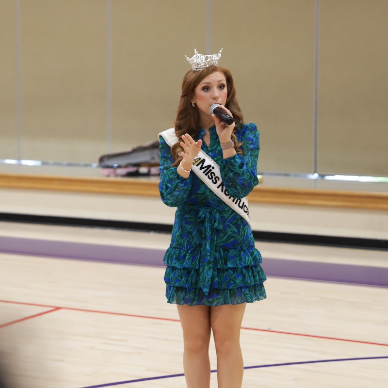 Chapel Tinius in a blue dress holding a microphone speaking in a school gym