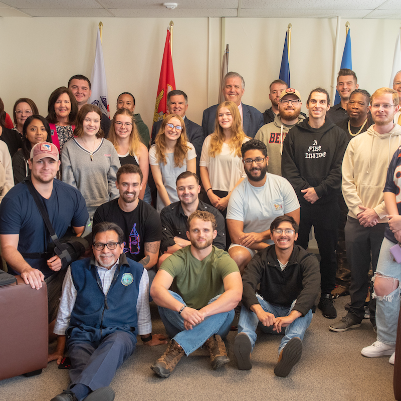 A large group photo of approximately 30 people of diverse ages, genders, and ethnicities in a room. They are arranged in rows, with some sitting on couches in the foreground and others standing behind. The room in the Bruin Vet Center meeting space with flags displayed on the wall in the background.